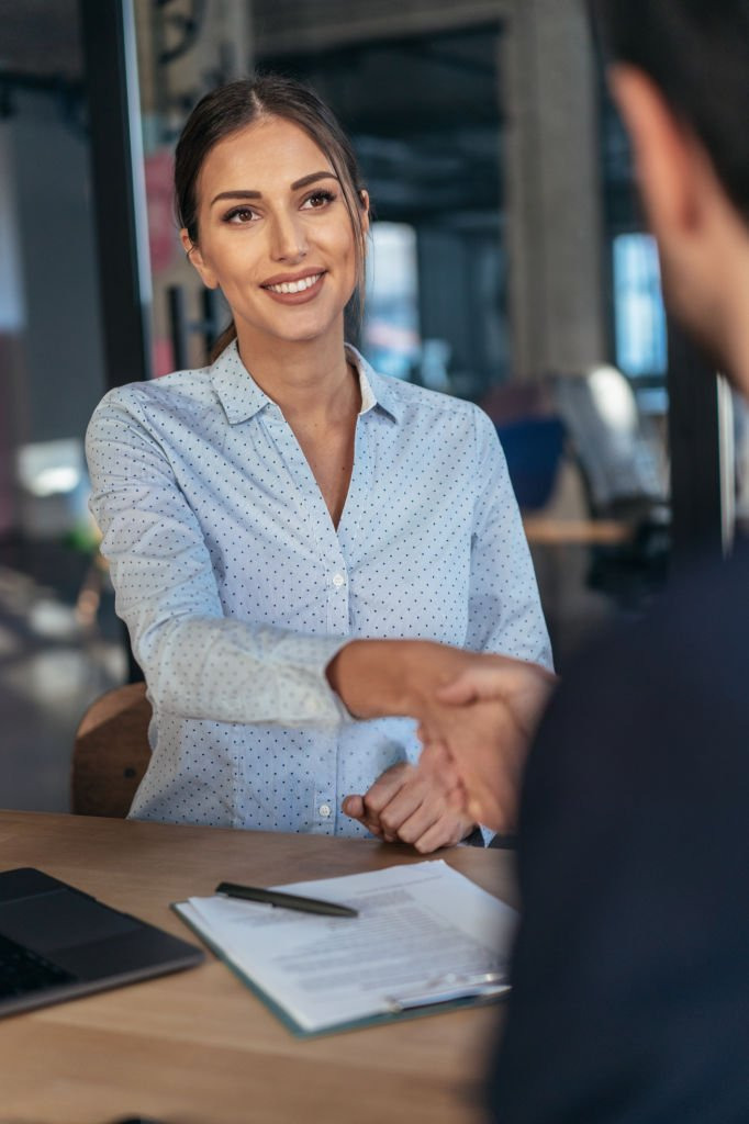 A smiling woman sitting at a desk shaking hands with a man over a signed contract.